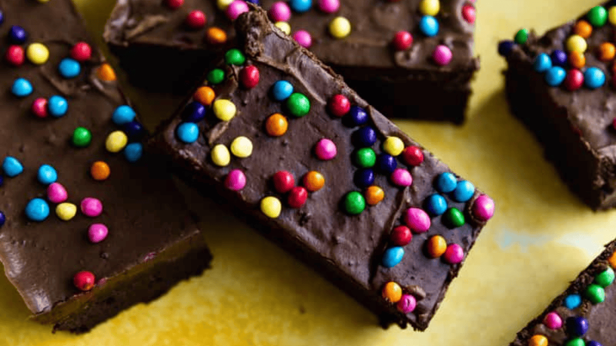 Rectangular chocolate brownies, perfect as 8x8 pan desserts, are topped with colorful round candy sprinkles and arranged on a yellow surface.