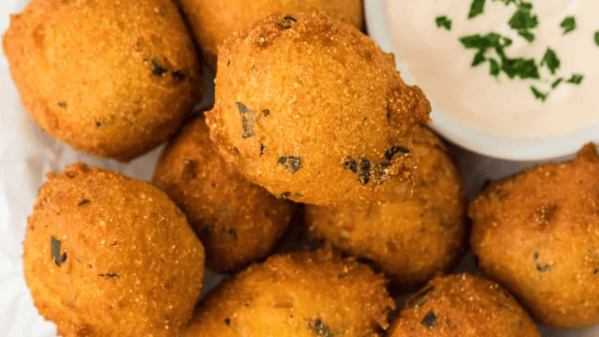 A close-up of golden-brown hush puppies arranged on a plate, served with a small bowl of creamy white dipping sauce garnished with chopped herbs—classic old school party snacks perfect for sharing.