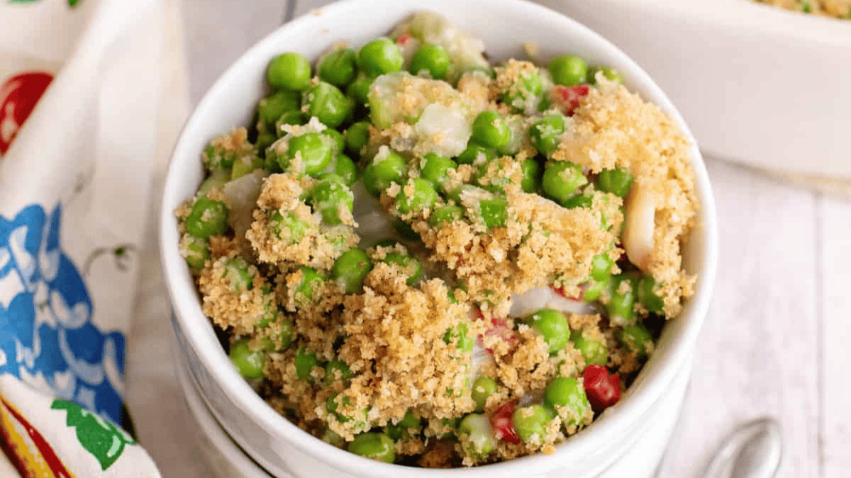 A white bowl filled with a baked casserole featuring green peas, chopped vegetables, a creamy sauce, and a golden breadcrumb topping—perfect for spring recipes.