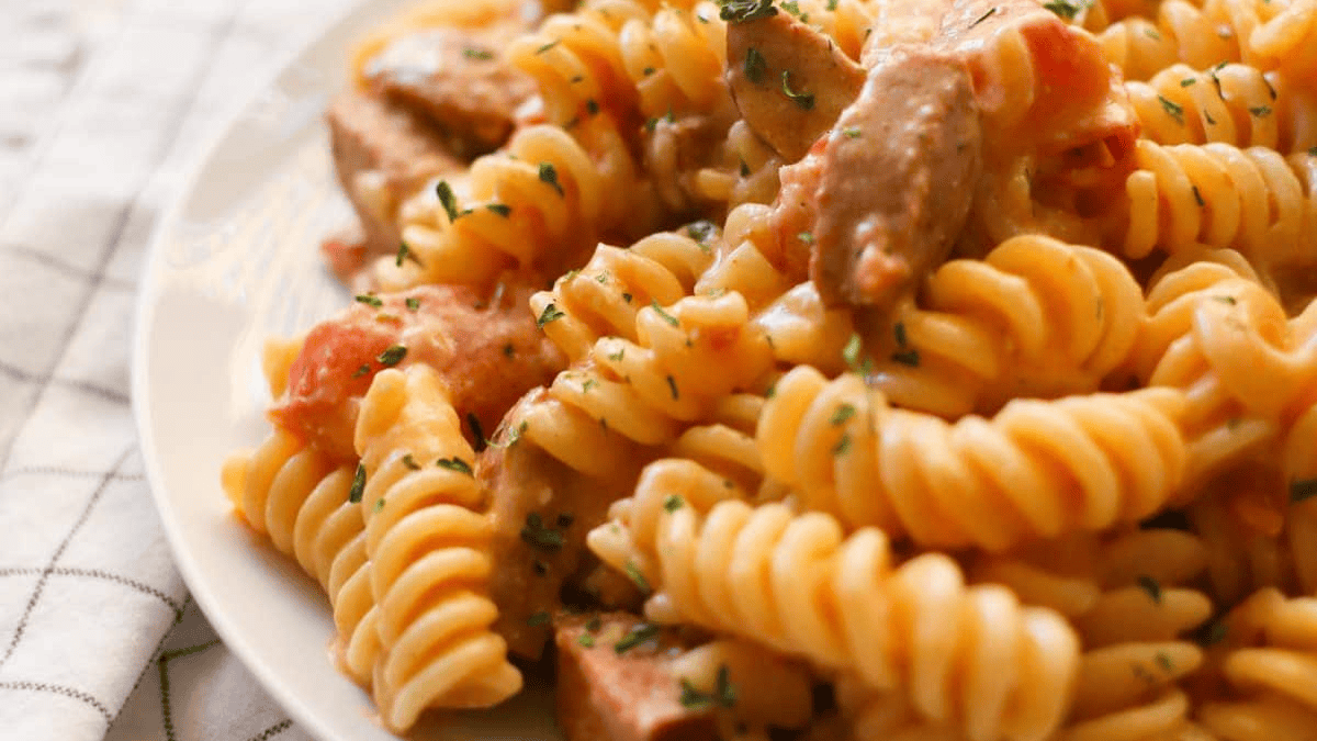 Close-up of creamy rotini pasta with pieces of meat and a zesty tomato-based sauce, inspired by cajun recipes, garnished with chopped herbs and served on a white plate.