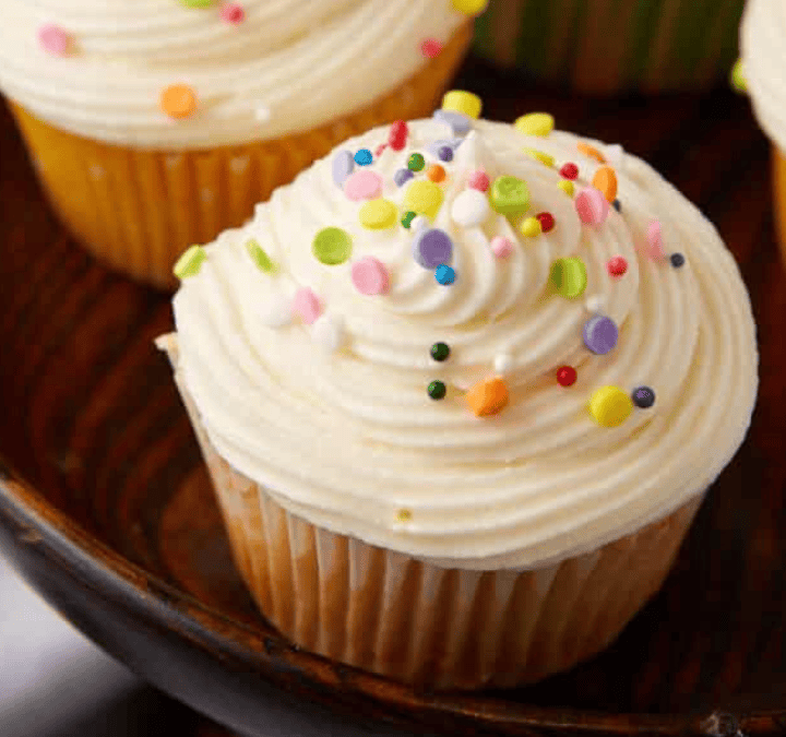 Four cupcakes with white frosting and sprinkles on a wooden plate.
