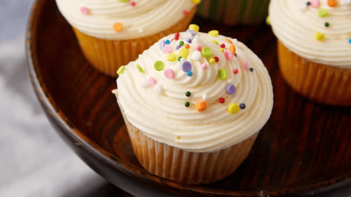 Four cupcakes with white frosting and sprinkles on a wooden plate.