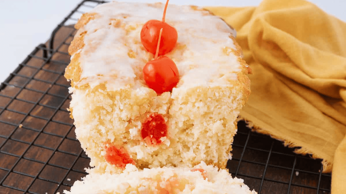 A glazed loaf cake with chopped red cherries inside and three whole cherries on top, placed on a cooling rack next to a yellow cloth—a delightful choice for sweet Easter breakfast recipes.