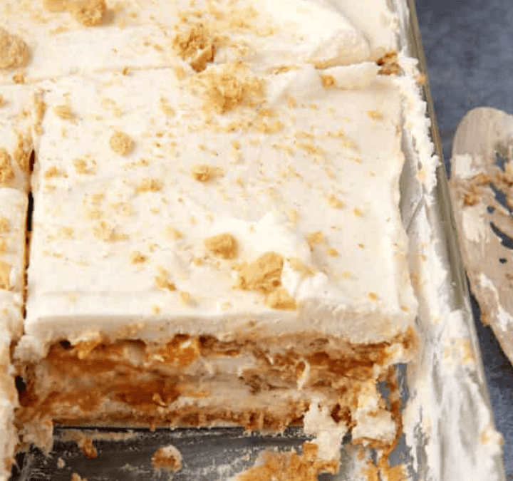 A close-up of a layered dessert with creamy white layers and crumbled cookies or crackers on top, partially sliced in a glass dish, with a serving spatula and forks nearby.