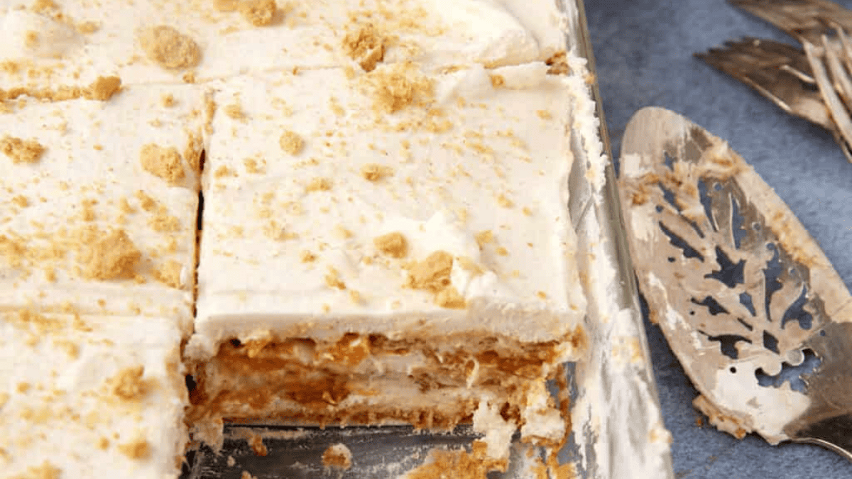 A close-up of a layered dessert with creamy white layers and crumbled cookies or crackers on top, partially sliced in a glass dish, with a serving spatula and forks nearby.
