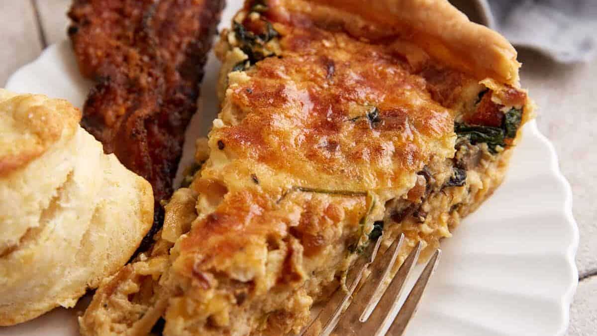A slice of spinach quiche on a plate with a biscuit and bacon.