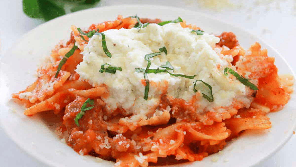 A plate of bowtie pasta with tomato sauce, ground meat, a generous dollop of creamy ricotta cheese, and fresh basil strips sprinkled on top—perfect for those craving delightful spring pastas.