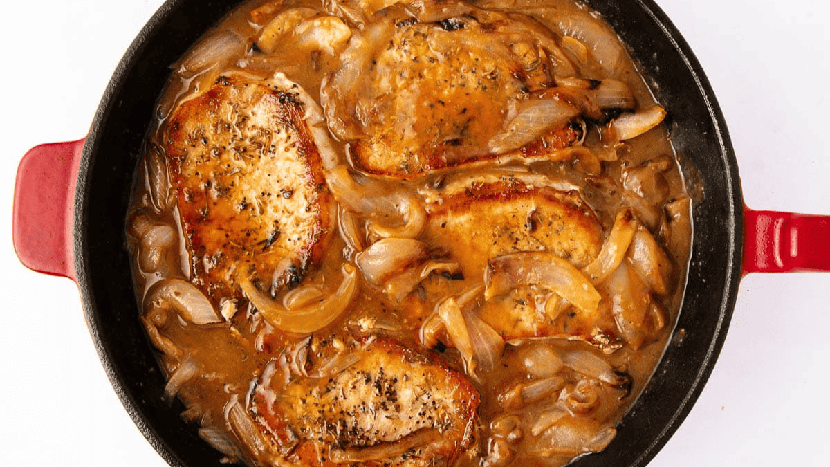 Three seasoned pork chops simmer with sliced onions and rich brown gravy, creating a hearty and savory dish—perfect for easy meals. The skillet, with red handles, sits on a white surface.