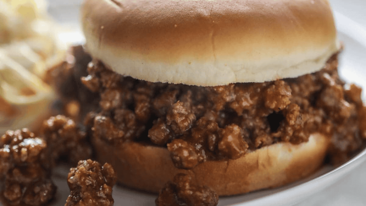 A close-up of a sloppy joe sandwich with a soft hamburger bun and seasoned ground beef spilling out, served on a white plate—an old fashioned dinner classic.