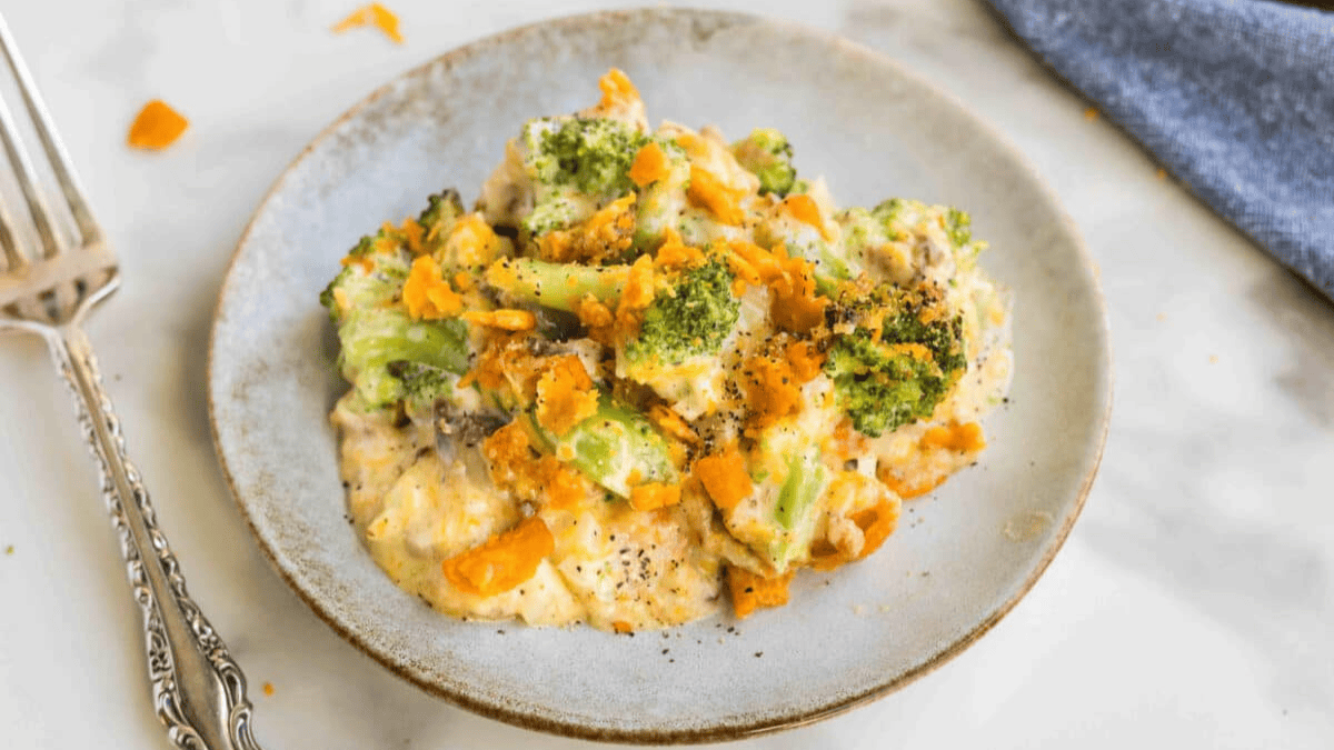 A plate of creamy broccoli casserole, a classic from old school meals, topped with melted cheese and crushed crackers, served on a light grey plate with a fork beside it on a white surface.