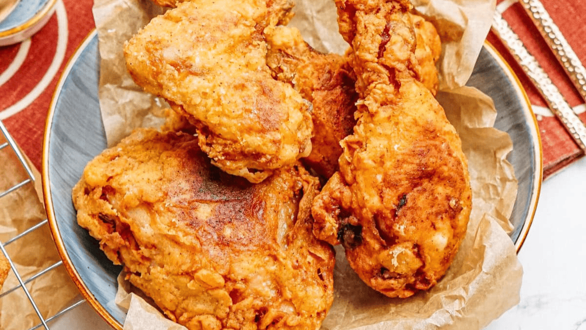 A bowl lined with parchment paper holds several pieces of crispy, golden-brown fried chicken, reminiscent of old school southern recipes, placed on a white surface near a red and gold napkin.
