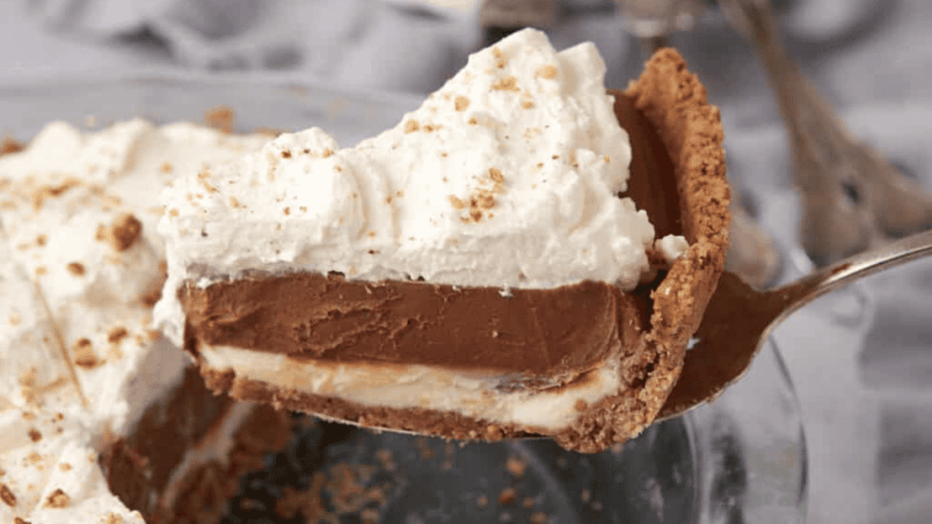 A close-up of a slice of chocolate cream pie, inspired by desserts around the world, with whipped cream on top, a rich chocolate filling layer, and a crumbly pie crust, being lifted from a glass pie dish.