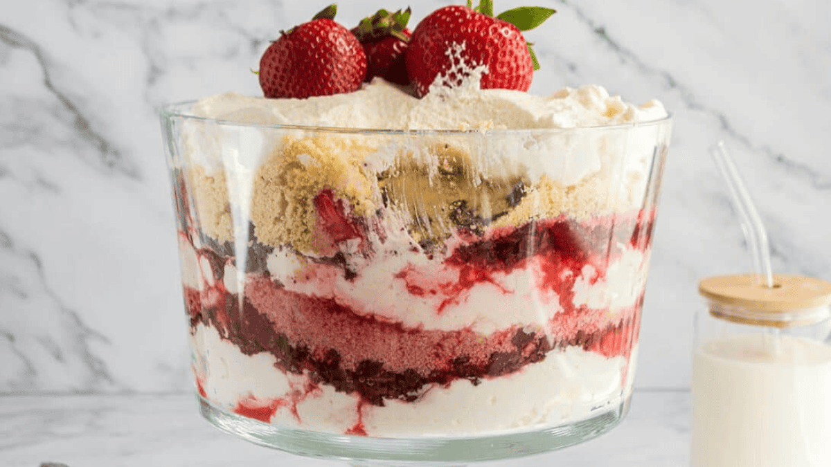 A glass trifle bowl showcases layered desserts with whipped cream, cake, berry sauce, crumbled cookies, and fresh strawberries against a marble background. A glass of milk with a straw sits beside it.