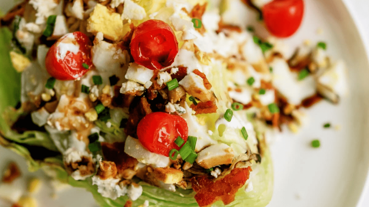A close-up of a wedge salad topped with chopped hard-boiled eggs, cherry tomatoes, bacon bits, crumbled cheese, and chives on a white plate—perfect for serving as one of your Easter sides.