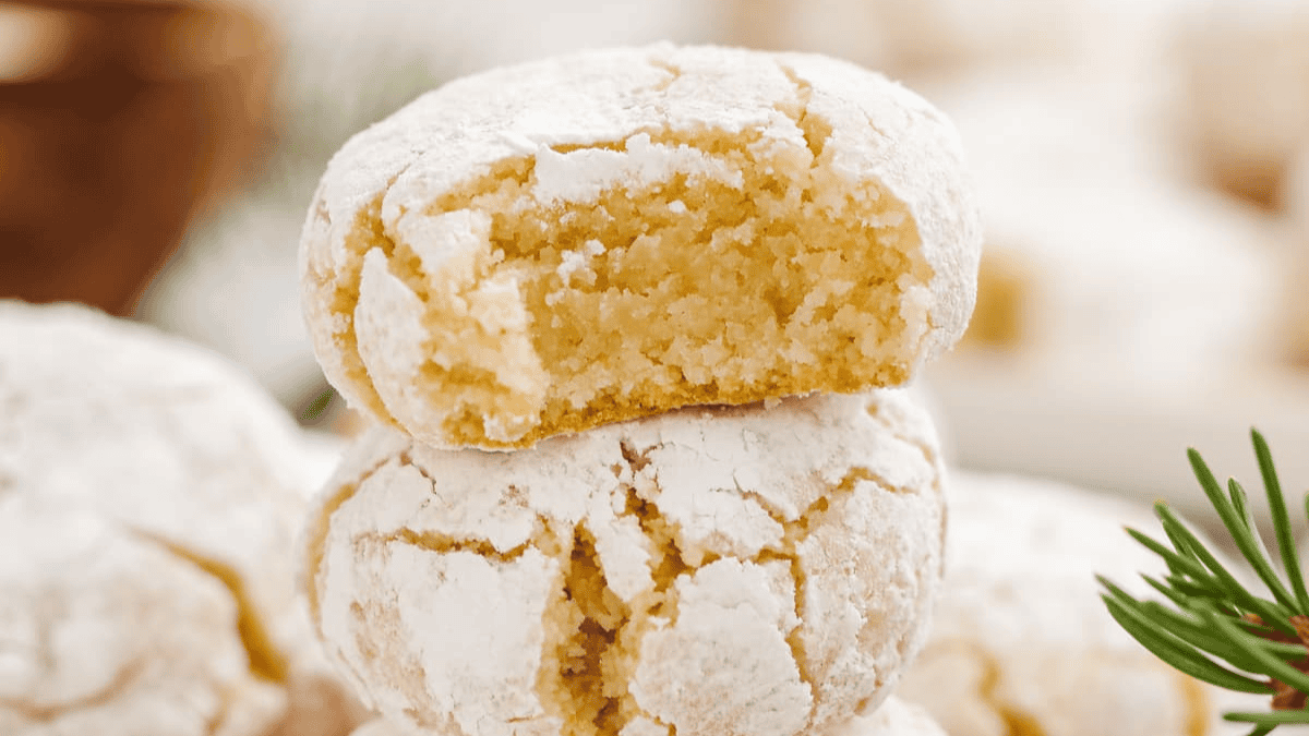 Two round, cracked Easter cookies dusted with powdered sugar; one is stacked on top of the other and has a bite taken out, revealing a soft, chewy inside. A sprig of rosemary is visible in the corner.