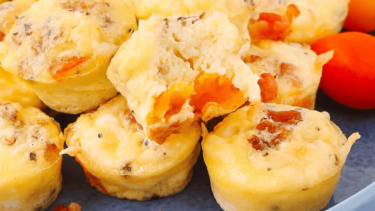 A close-up of egg muffins with visible bits of bacon and cheese, stacked on a plate. One muffin is split open, revealing its soft, cheesy interior. Perfect for leftover egg recipes, bright cherry tomatoes are placed nearby.