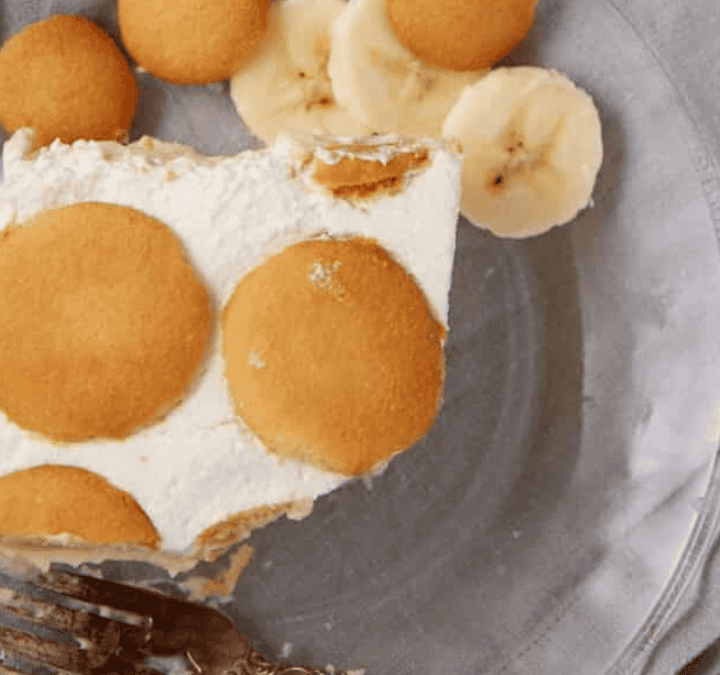 A slice of banana pudding, reminiscent of spring cakes, topped with round vanilla wafers and banana slices on a clear glass plate, with a fork and a light gray napkin beside it.