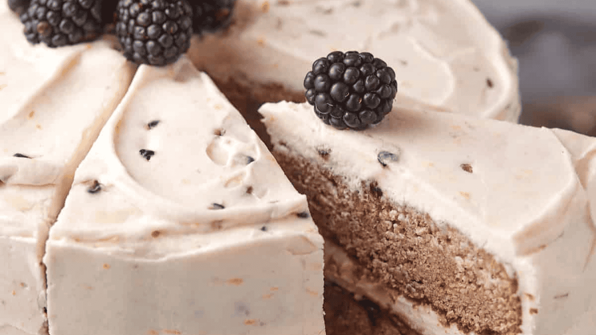 A frosted spring cake is topped with blackberries. One slice is being pulled out, revealing the cake&rsquo;s light brown interior and creamy white frosting with dark specks.