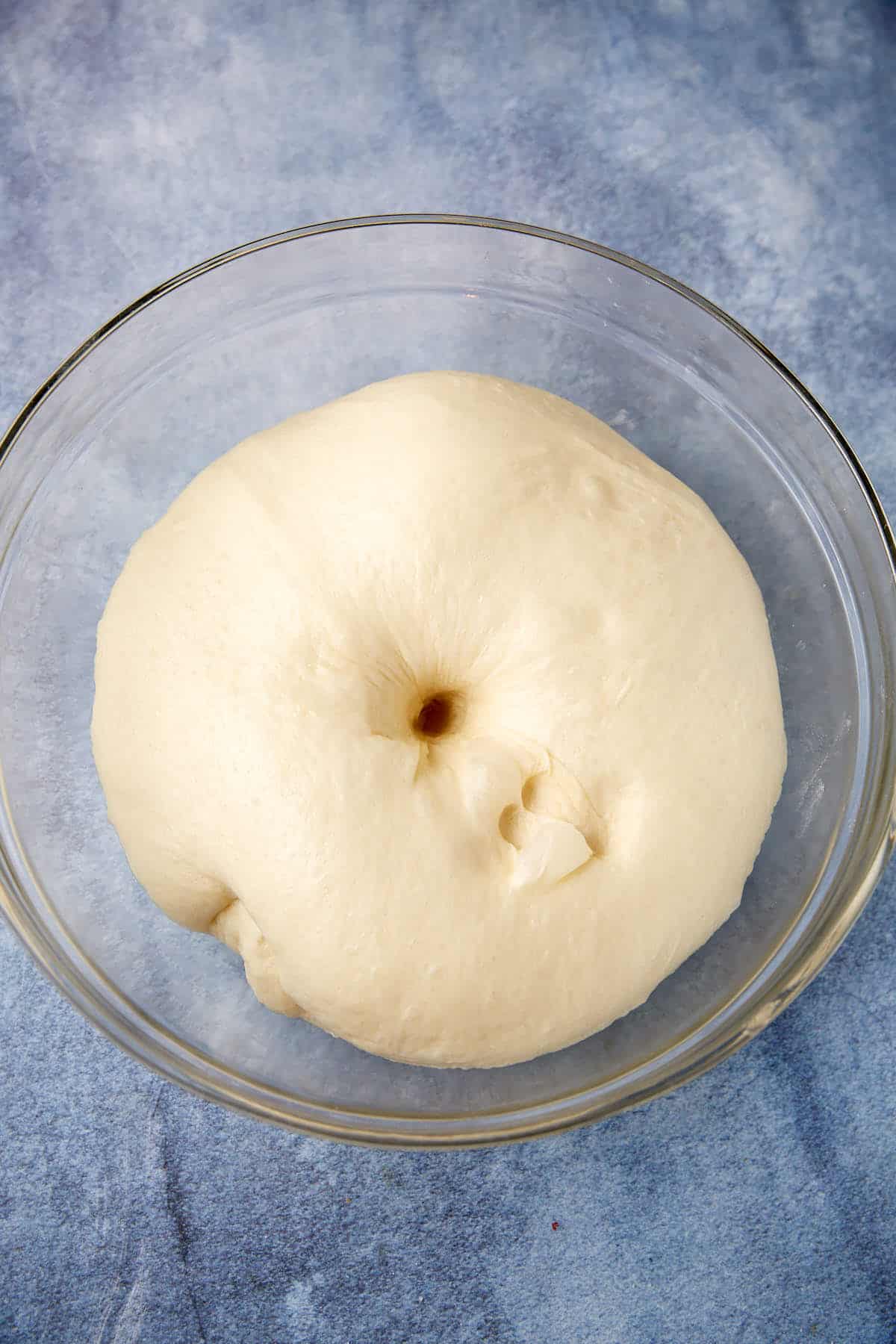 risen buttermilk yeast roll dough with a hole poked into it resting in a glass bowl.