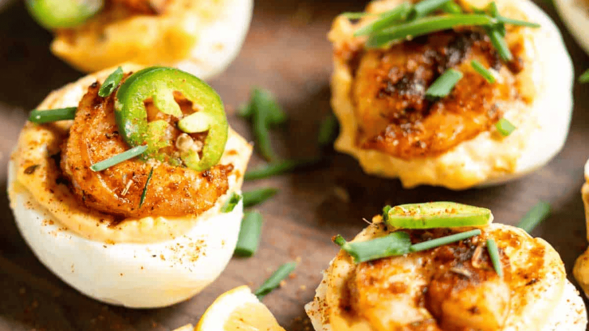 Deviled eggs topped with seasoned shrimp, sliced jalapeños, and chopped chives are artfully arranged on a wooden surface.