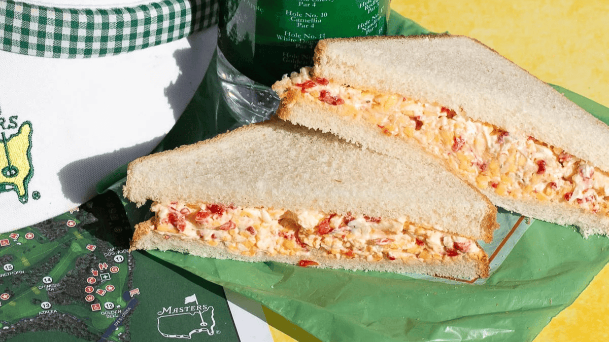 A classic pimento cheese sandwich on white bread, cut in half and placed on green wax paper, sits next to a Masters logo cooler, water bottle, and part of an event map—perfect national picnic day finger foods on a yellow surface.