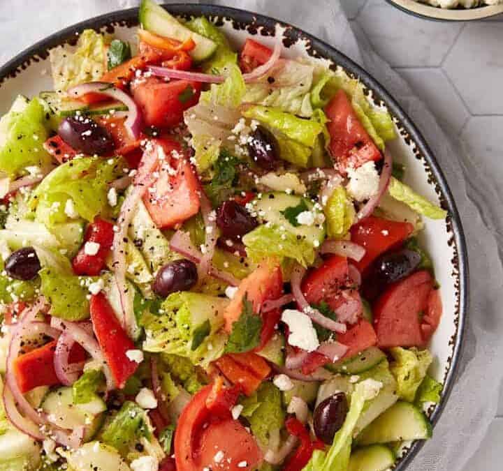 A bowl of Greek salad with lettuce, tomatoes, cucumbers, red onions, Kalamata olives, and crumbled feta cheese on a white surface—perfect for spring recipes. Small bowls of olives and feta are nearby.