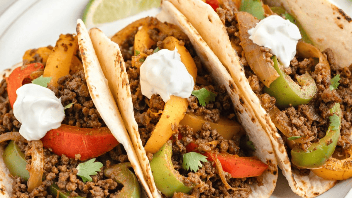 Three soft tacos filled with seasoned ground beef, sautéed red, yellow, and green bell peppers, onions, and topped with dollops of sour cream, garnished with cilantro. Perfect for your next fiesta or as a tasty addition to Cinco de Mayo recipes. A lime wedge is visible in the background.