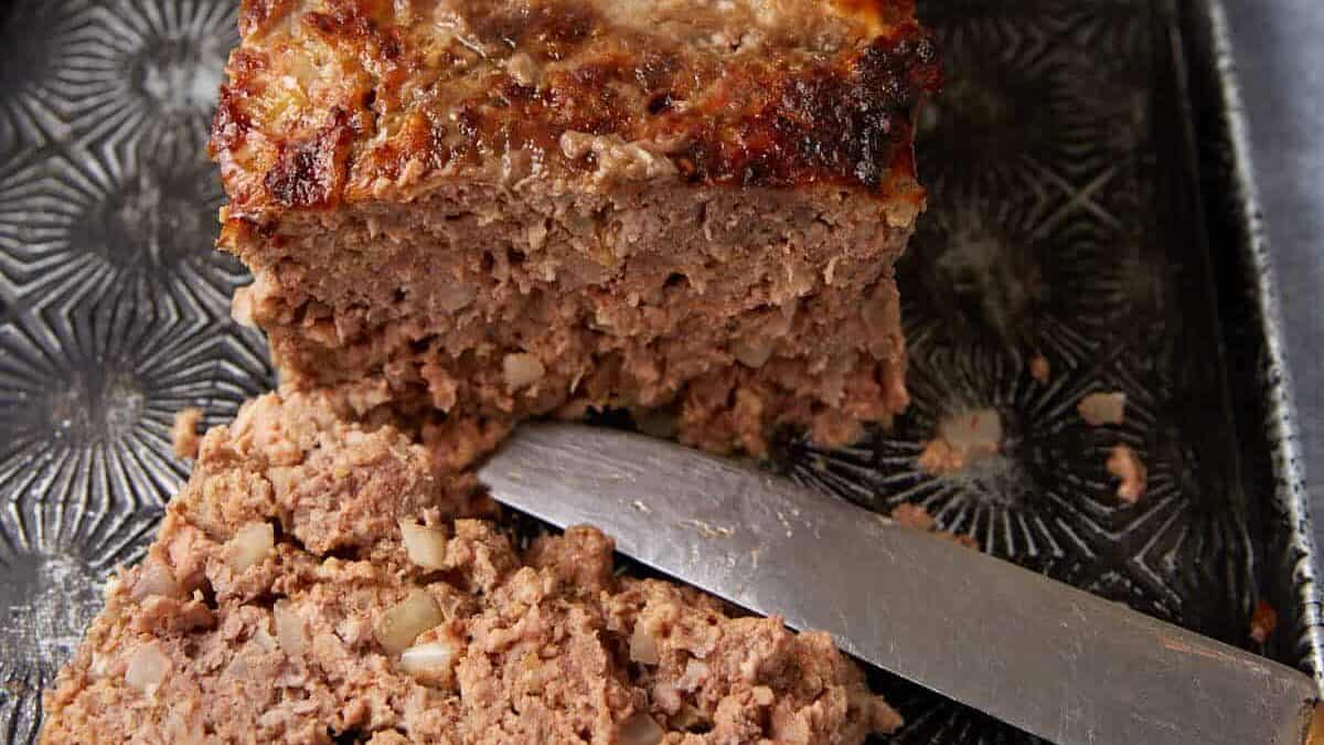 A well-cooked meatloaf sits on a textured, rectangular tray. One slice has been cut from the decadent creation, and a silver knife lies next to it. The meatloaf appears moist and has a browned, crusty top. A small container in the background holds a red sauce or ketchup for serving.