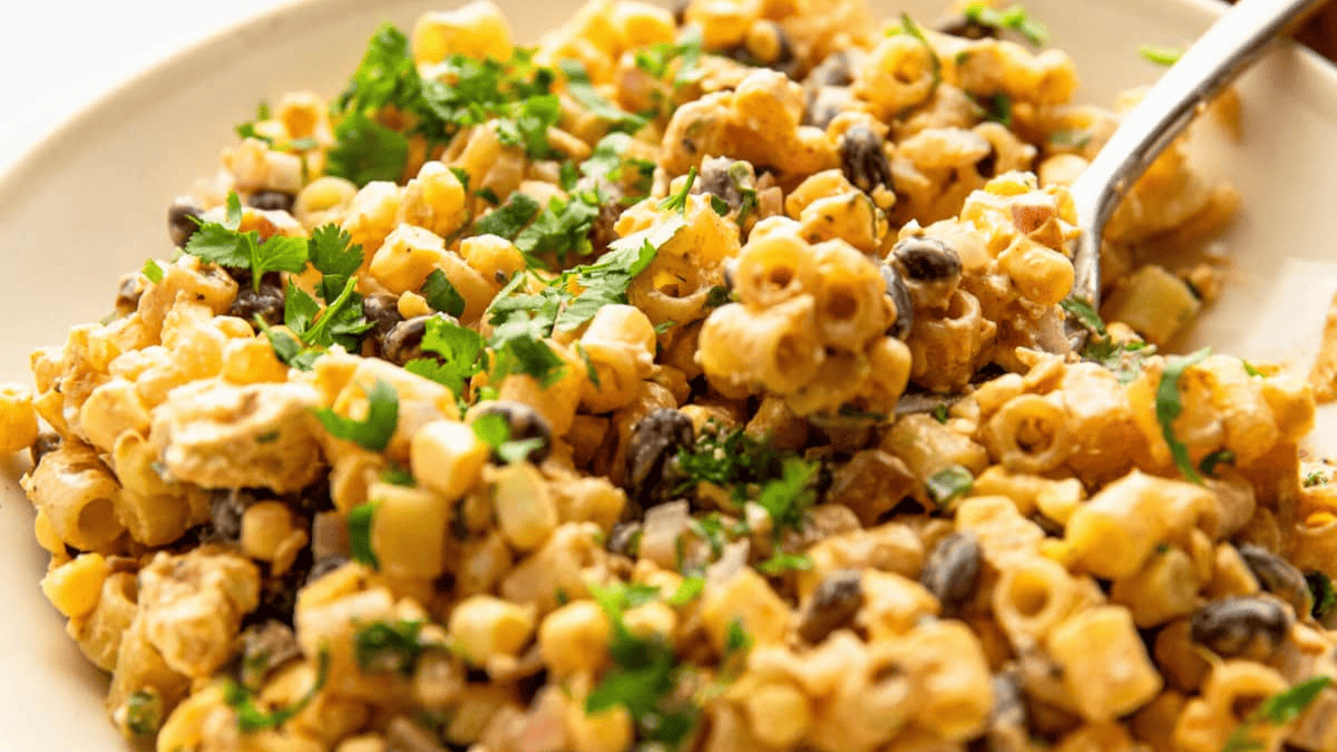 A close-up of a creamy pasta salad with black beans, corn, chopped onion, and fresh cilantro on a white plate, served with a spoon—perfect for adding to your list of Cinco de Mayo recipes.