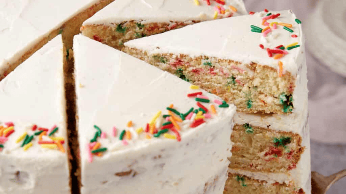 A close-up of a frosted layer cake with colorful rainbow sprinkles on top and inside, several slices cut—an irresistible choice for Mother’s Day desserts, with one slice slightly pulled out to reveal the festive interior.