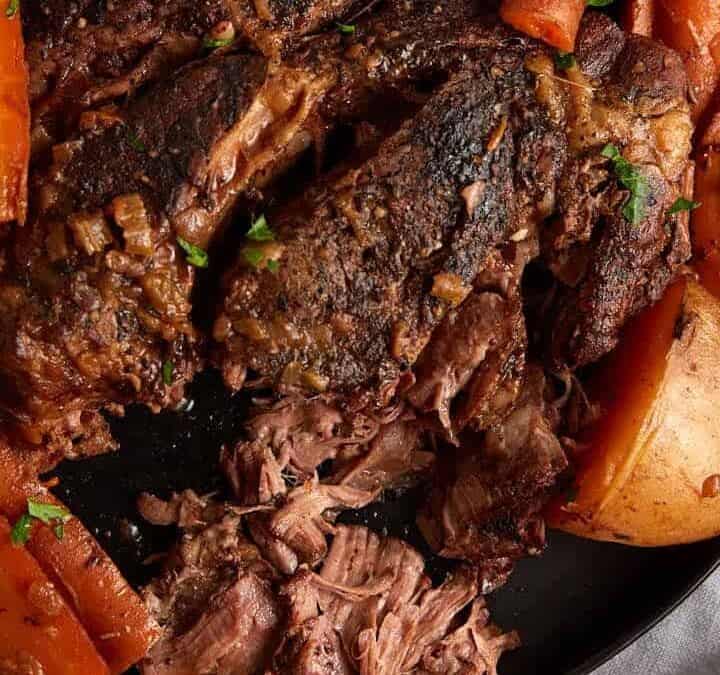 A large platter with a slow cooked roast beef surrounded by carrots and potatoes.