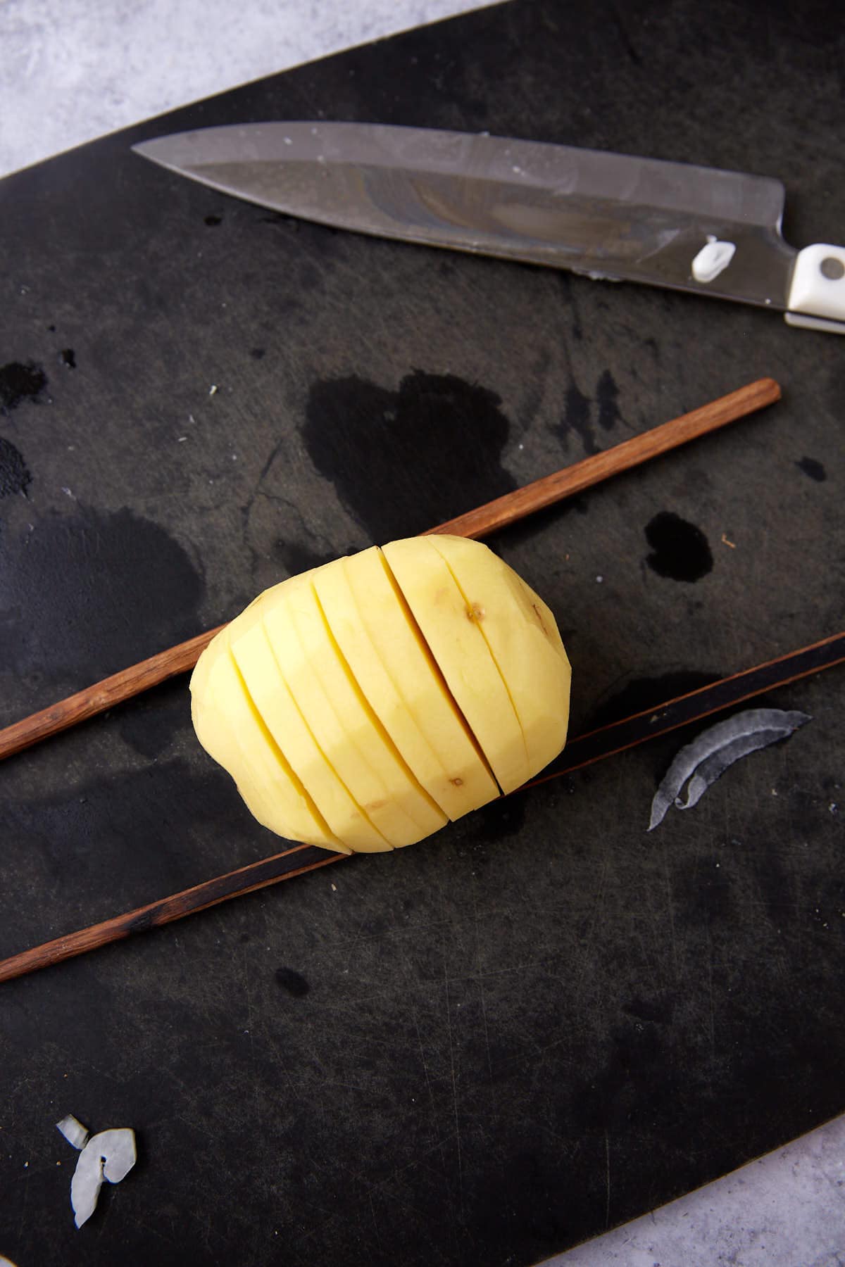 Yukon gold potato in between two chopsticks with slits in the potato and a knife in the background.