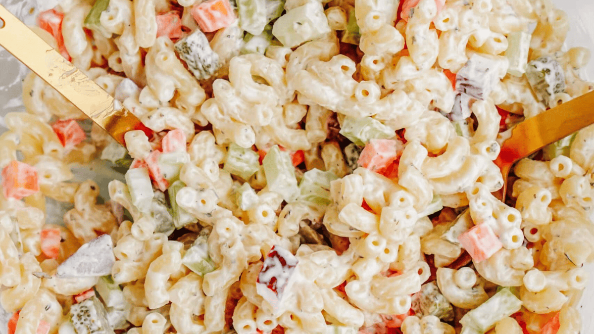 Close-up of creamy macaroni salad with elbow pasta, diced carrots, celery, pickles, and a mayonnaise-based dressing—just like recipes mom used to make—mixed together with two gold-colored utensils.