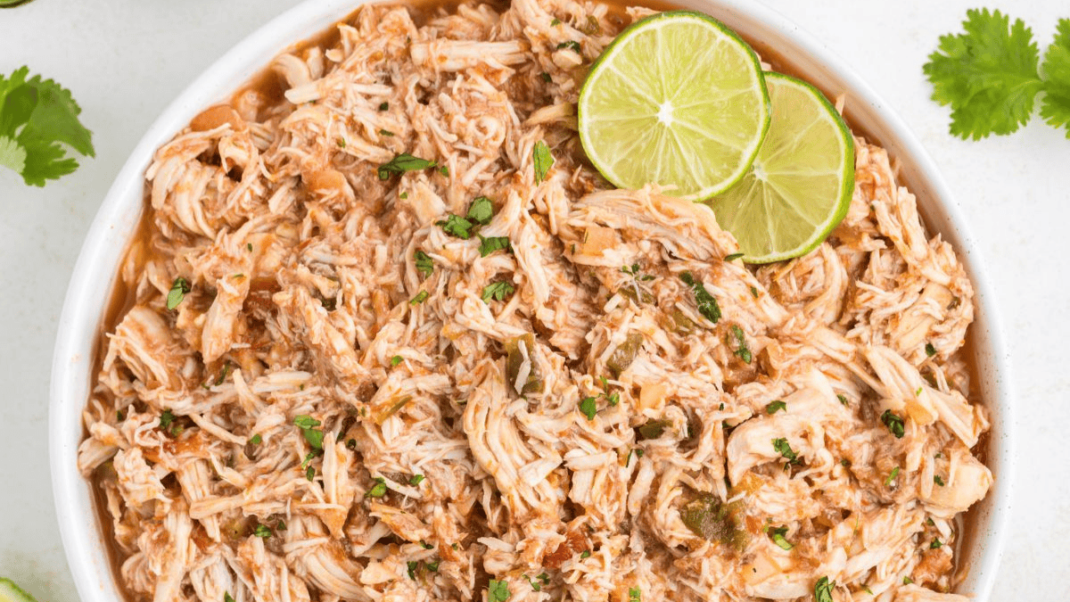 A bowl of shredded chicken garnished with chopped herbs and two slices of lime, with cilantro leaves nearby on a white background—perfect for adding to your list of Cinco de Mayo recipes.