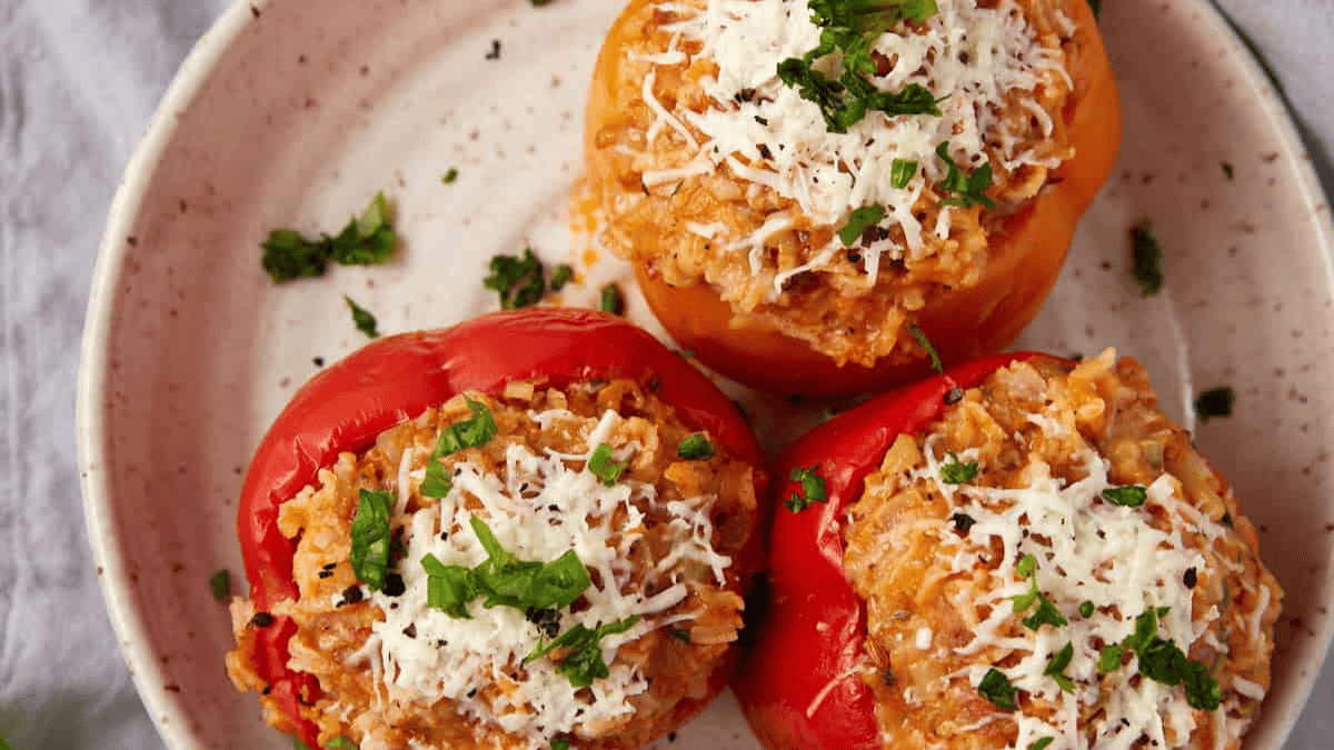 Three stuffed bell peppers, inspired by classic 1980s recipes, two red and one orange, filled with rice and topped with shredded cheese and chopped fresh herbs, served on a light-colored plate.