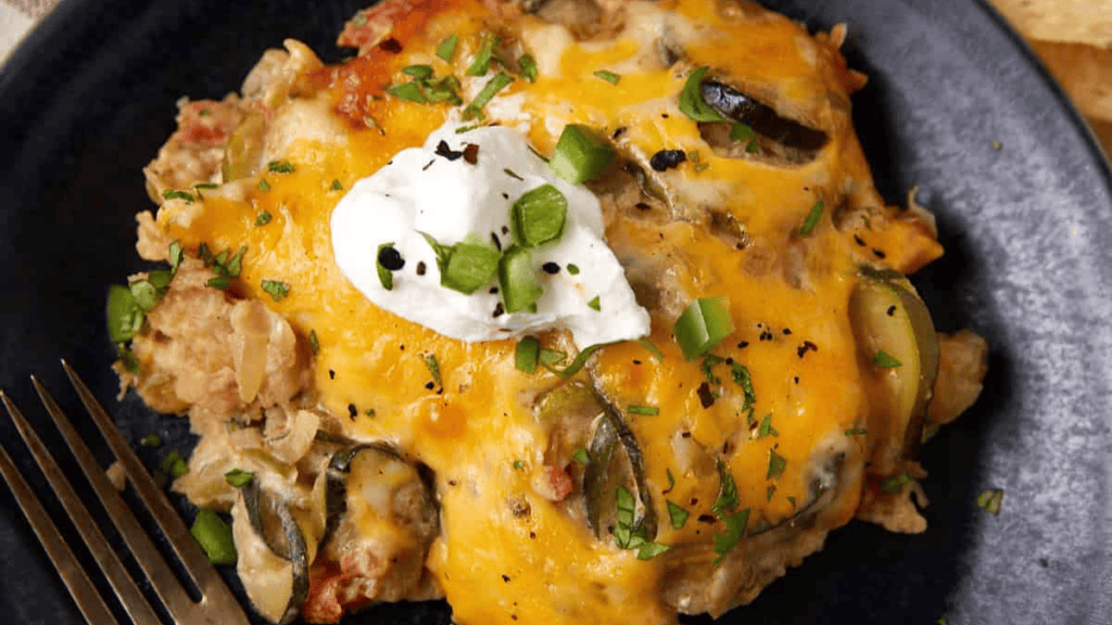 A serving of cheesy casserole topped with sour cream and chopped green onions sits on a dark plate next to a fork. The dish, ideal for Cinco de Mayo recipes, features melted cheddar cheese and visible slices of jalapeño.