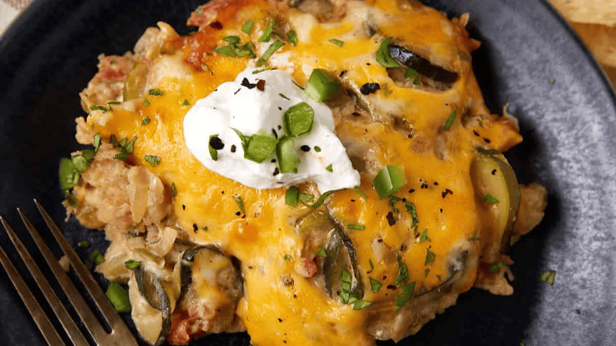 A serving of cheesy casserole topped with sour cream and chopped green onions sits on a dark plate next to a fork. The dish, ideal for Cinco de Mayo recipes, features melted cheddar cheese and visible slices of jalapeño.
