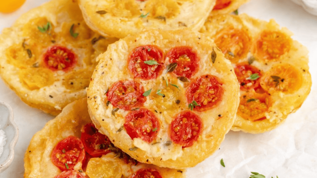 Round mini flatbreads topped with red and yellow cherry tomato slices, fresh herbs, and seasoning—perfect finger foods for celebrating National Picnic Day, arranged beautifully on a white surface.