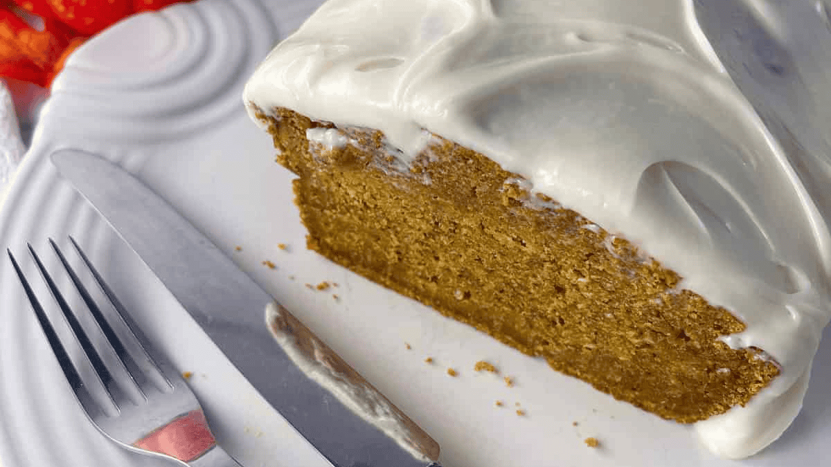 A close-up of a thick slice of pumpkin bread topped with creamy white frosting on a white plate, with a fork and knife placed beside it.
