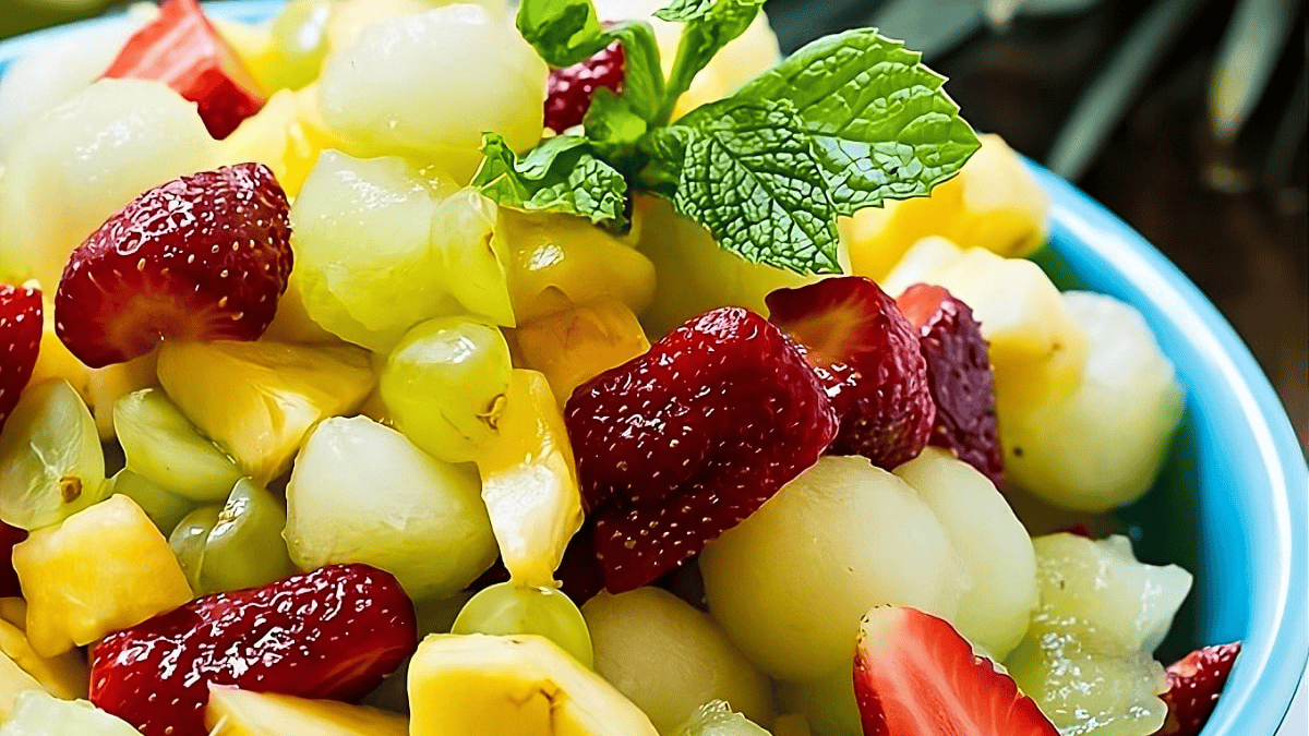 A colorful fruit salad with strawberries, melon balls, pineapple chunks, and grapes in a blue bowl, garnished with a fresh mint sprig.