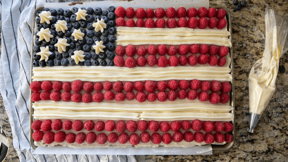 A sheet cake decorated like the American flag with blueberries and star-shaped white frosting for the stars, and rows of raspberries and white frosting for the stripes, on a granite countertop.