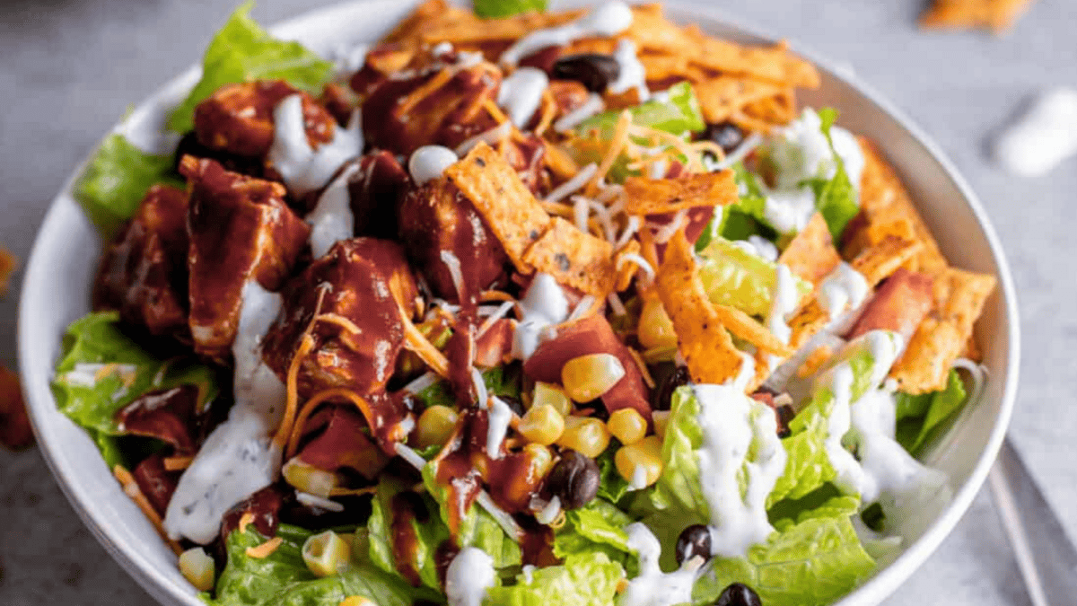 A bowl of salad topped with grilled chicken, black beans, corn, shredded cheese, tortilla strips, and drizzled with creamy dressing and barbecue sauce.