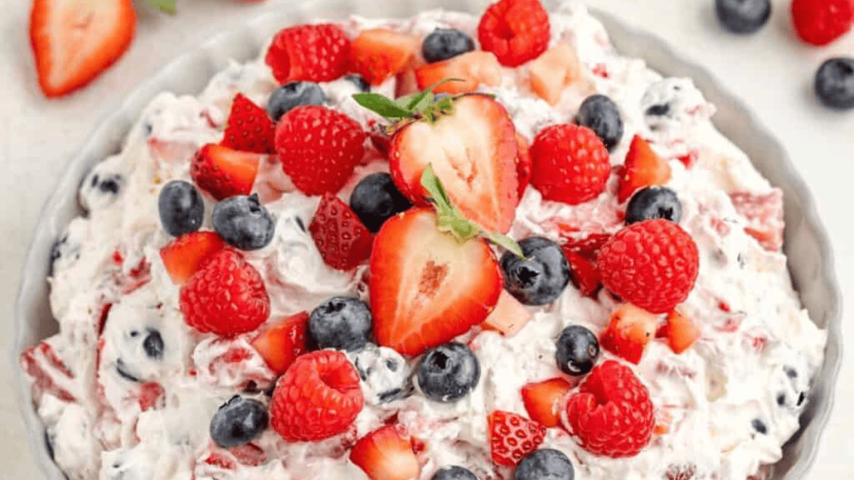 A bowl of creamy fruit salad topped with sliced strawberries, blueberries, and raspberries. The salad is mixed with whipped cream and pieces of fruit, creating a colorful and textured dessert.