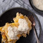 A serving of biscuits and gravy casserole topped with creamy sausage gravy, placed on a dark plate with a fork. A bowl of extra gravy sits nearby on a light gray cloth.