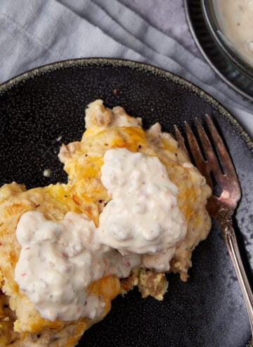 A serving of biscuits and gravy casserole topped with creamy sausage gravy, placed on a dark plate with a fork. A bowl of extra gravy sits nearby on a light gray cloth.