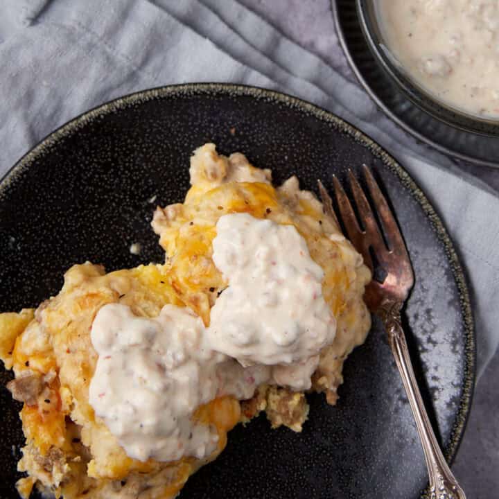 A serving of biscuits and gravy casserole topped with creamy sausage gravy, placed on a dark plate with a fork. A bowl of extra gravy sits nearby on a light gray cloth.