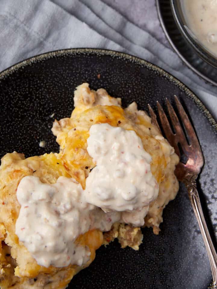 A serving of biscuits and gravy casserole topped with creamy sausage gravy, placed on a dark plate with a fork. A bowl of extra gravy sits nearby on a light gray cloth.