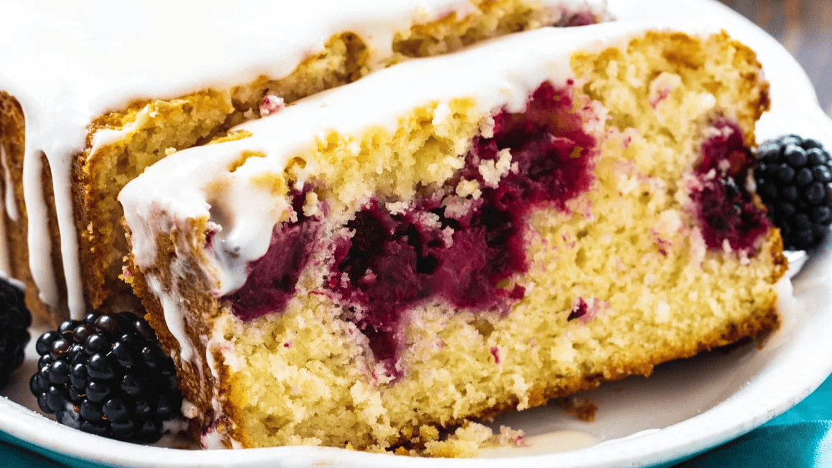 A close-up of a slice of moist blackberry loaf cake with white icing on top, showing a swirl of blackberries inside. Two fresh blackberries are on the plate beside the cake.