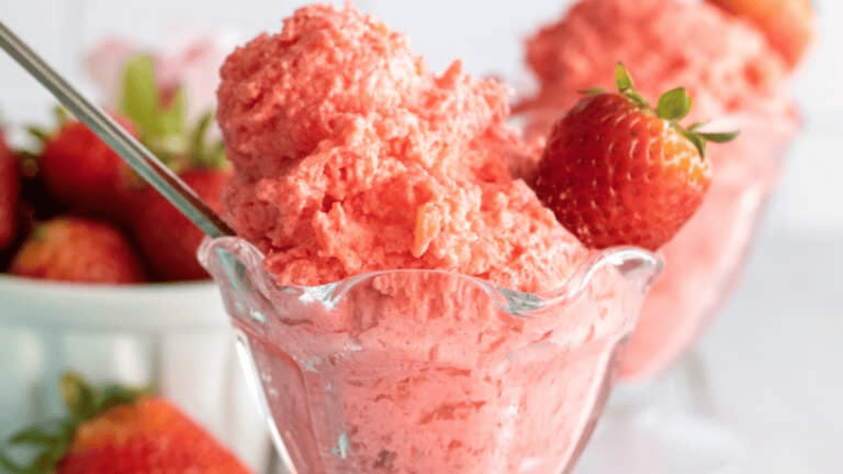A glass dessert dish filled with creamy, pink strawberry mousse, garnished with a whole fresh strawberry, with more strawberries visible in a bowl in the background.