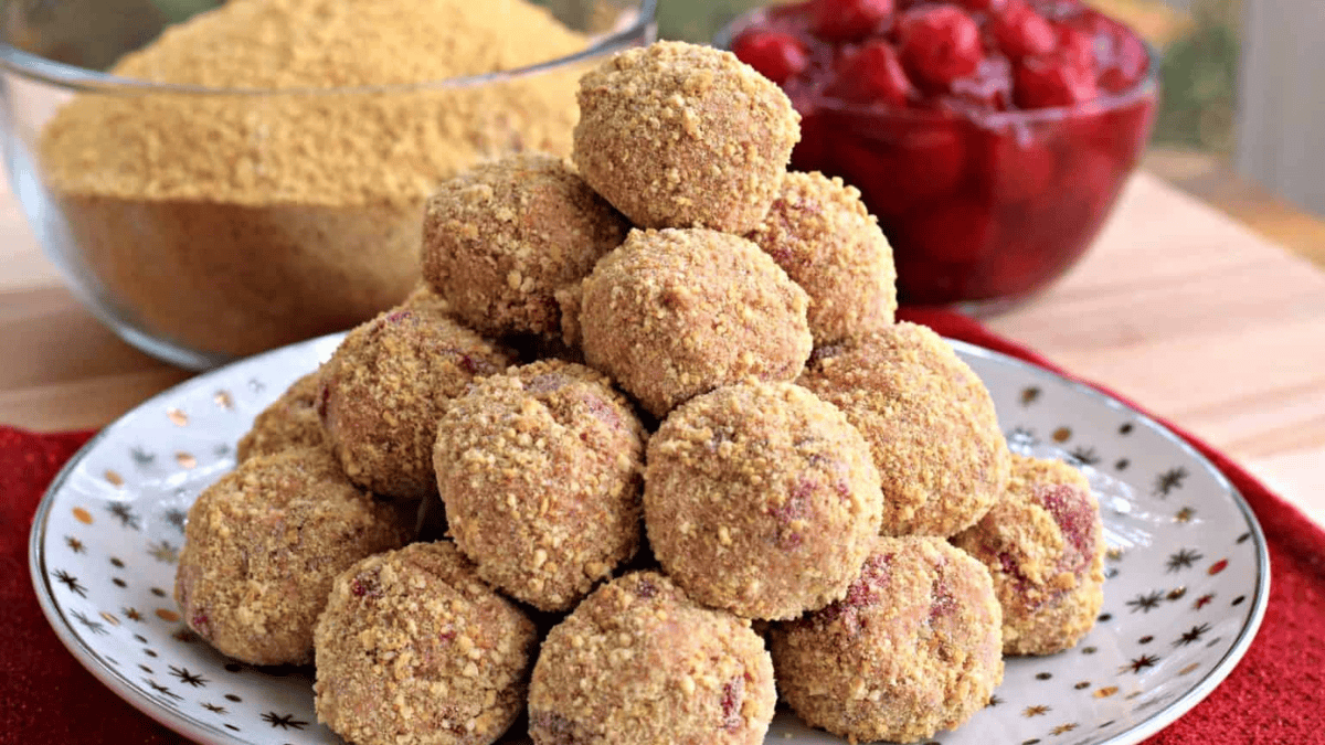 A plate stacked with round, crumb-coated dessert balls. In the background, there’s a bowl of crushed graham crackers and a bowl of bright red cherry filling. The plate sits on a wooden surface with a red napkin.